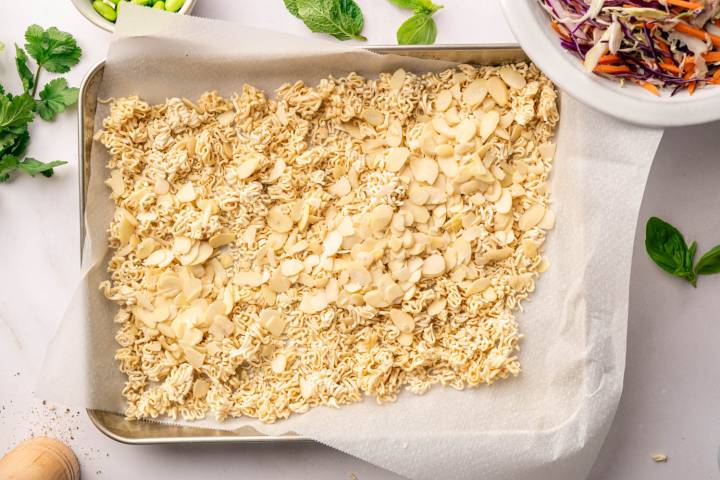 A baking sheet with crushed dry ramen noodles and sliced almonds on parchment paper, surrounded by fresh herbs and a bowl of colorful slaw, ready for preparation.